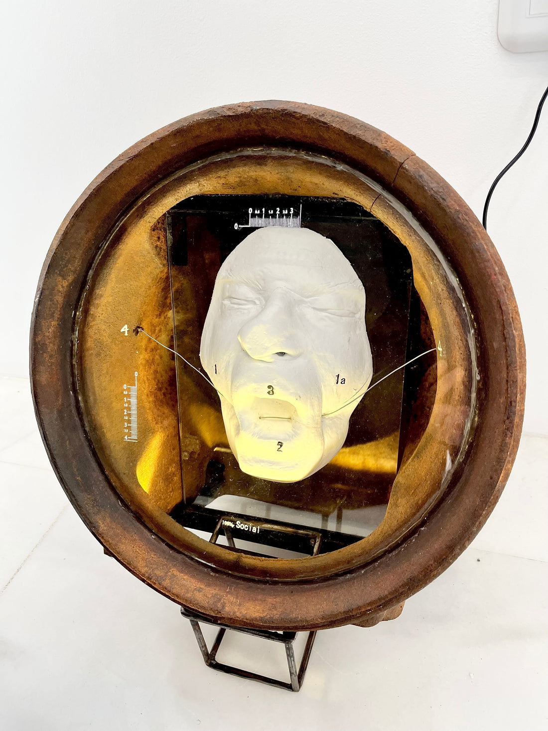 Plaster cast of a flattened human face housed inside a heavy, vintage cast-iron section of an Aga cooker, creating a visual resembling a 19th-century diver's helmet from the artwork 100% Social by Furst_Ori.