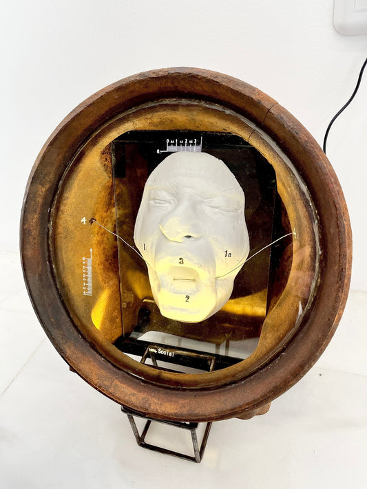 Plaster cast of a flattened human face housed inside a heavy, vintage cast-iron section of an Aga cooker, creating a visual resembling a 19th-century diver's helmet from the artwork 100% Social by Furst_Ori.