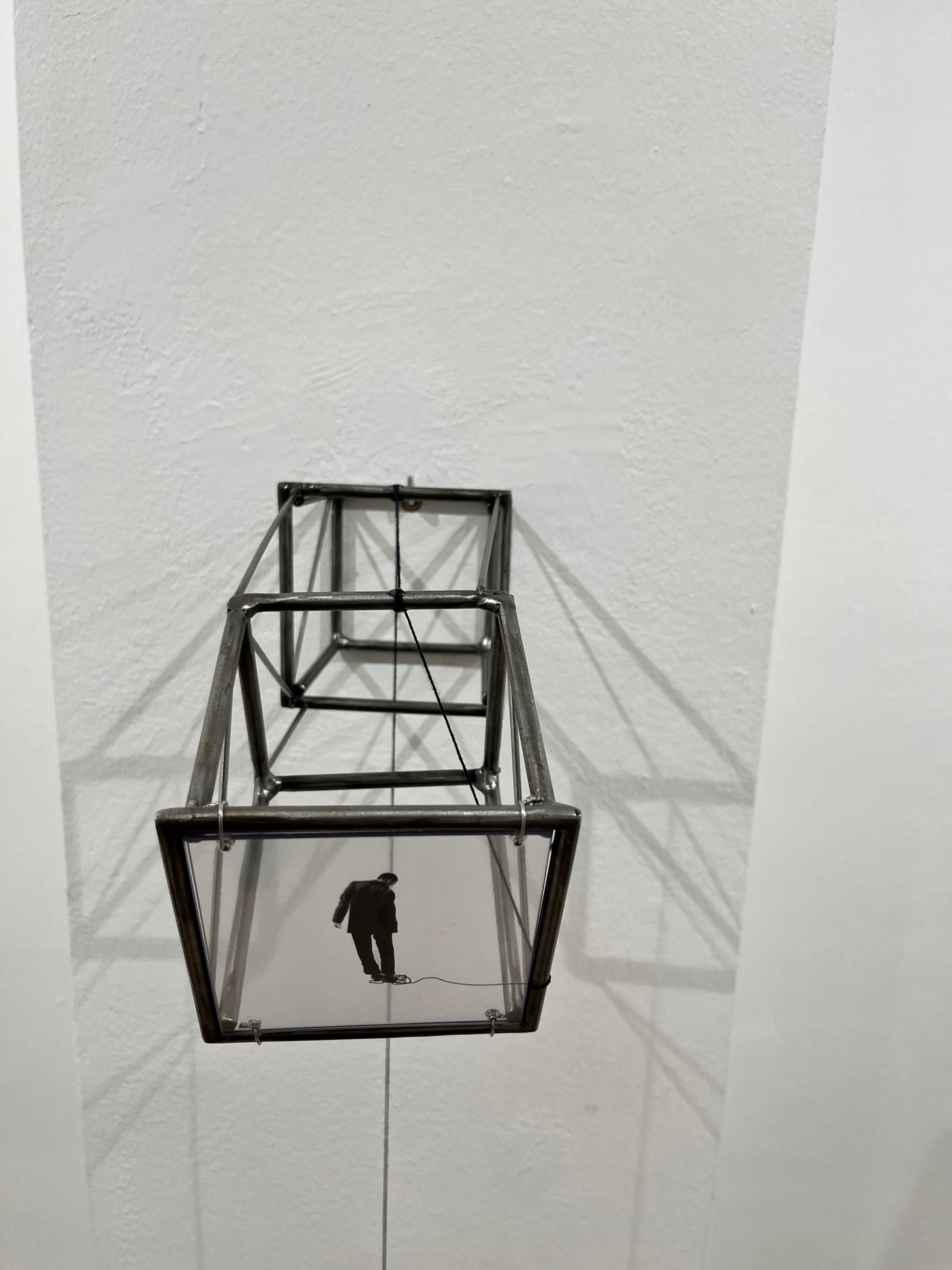 Hanging by a Thread is stylish, contemporary light and shadow sculpture, a geometric form given an asymetric twist, designed to lend any interior a quirky modernist elegance. An steel shadow artpiece depicting a man looking at his foot caught up in a thread leading to a stainless steel pendulum ball woven into the handmade structure. The image is a high quality print of an original Furst_Ori collage designed to caste a shadow within the frame.