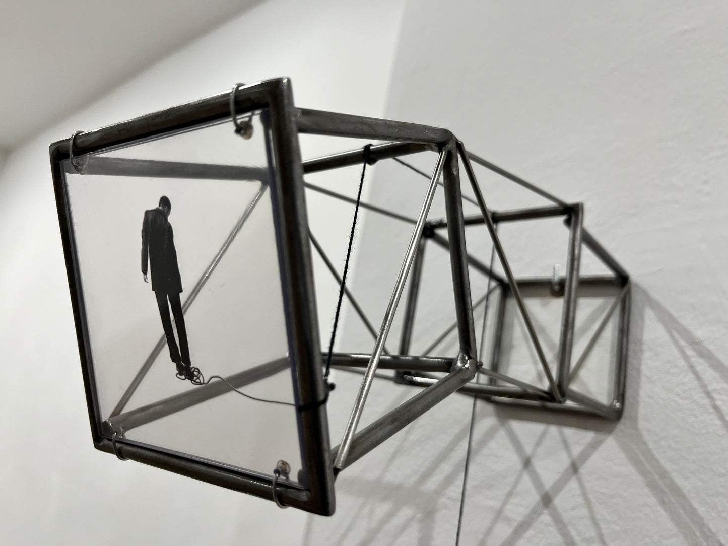 Hanging by a Thread is stylish, contemporary light and shadow sculpture, a geometric form given an asymetric twist, designed to lend any interior a quirky modernist elegance. An steel shadow artpiece depicting a man looking at his foot caught up in a thread leading to a stainless steel pendulum ball woven into the handmade structure. The image is a high quality print of an original Furst_Ori collage designed to caste a shadow within the frame.