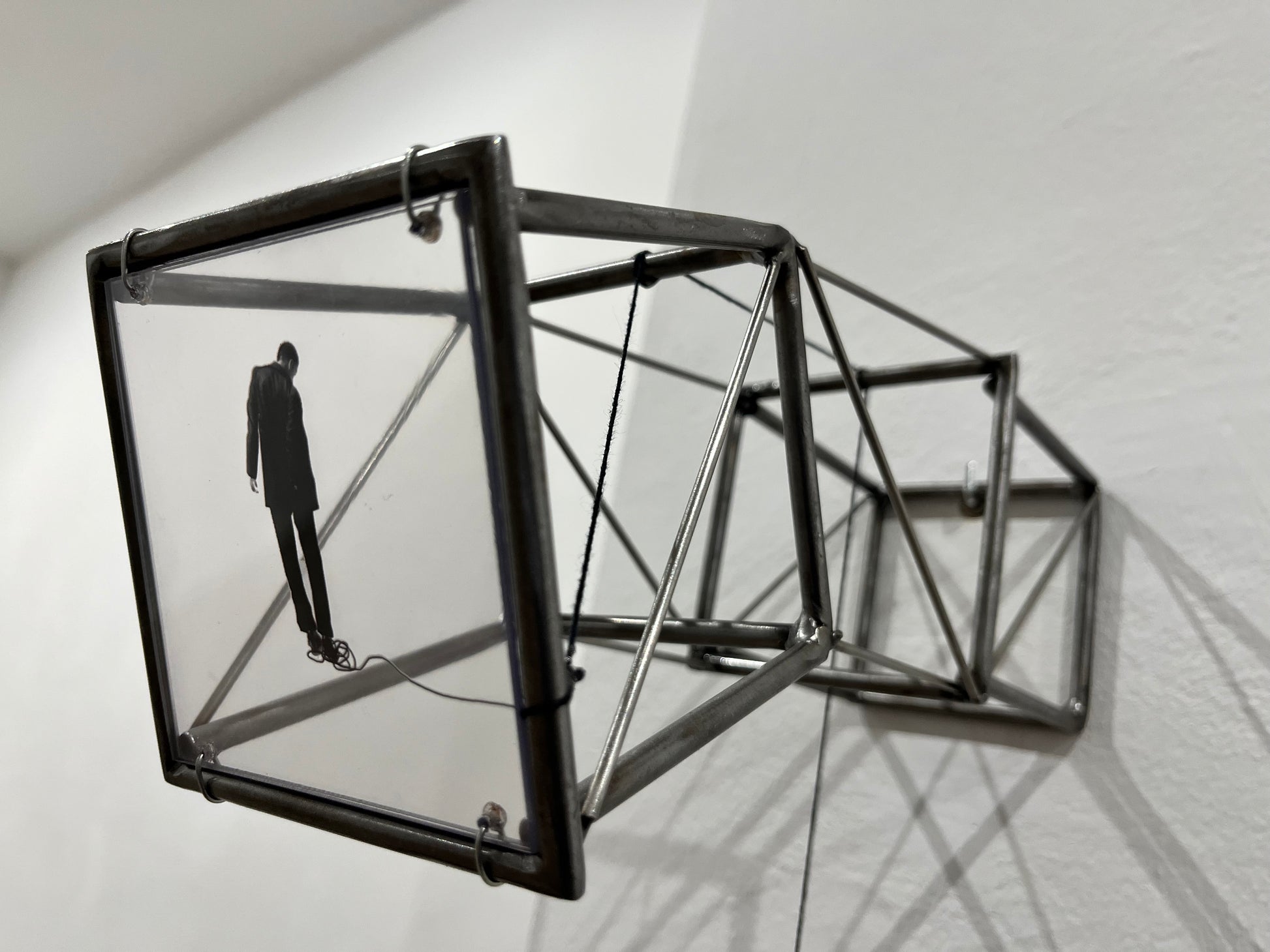 Hanging by a Thread is stylish, contemporary light and shadow sculpture, a geometric form given an asymetric twist, designed to lend any interior a quirky modernist elegance. An steel shadow artpiece depicting a man looking at his foot caught up in a thread leading to a stainless steel pendulum ball woven into the handmade structure. The image is a high quality print of an original Furst_Ori collage designed to caste a shadow within the frame.