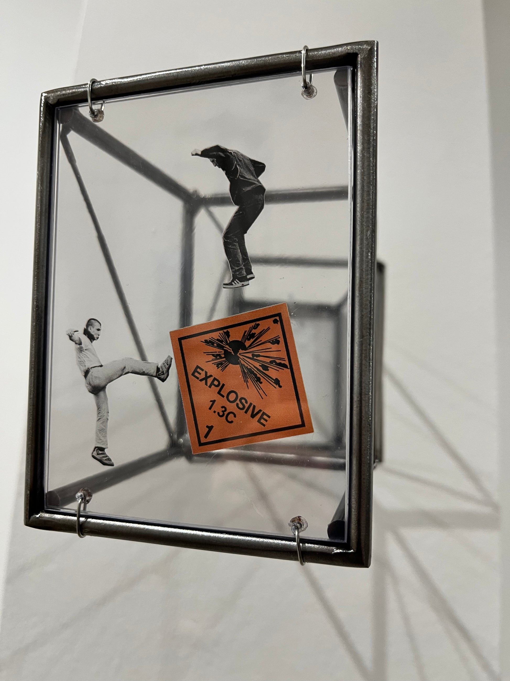 Artistic depiction of a person in a cube with an 'Explosive' label, framed against a white wall.