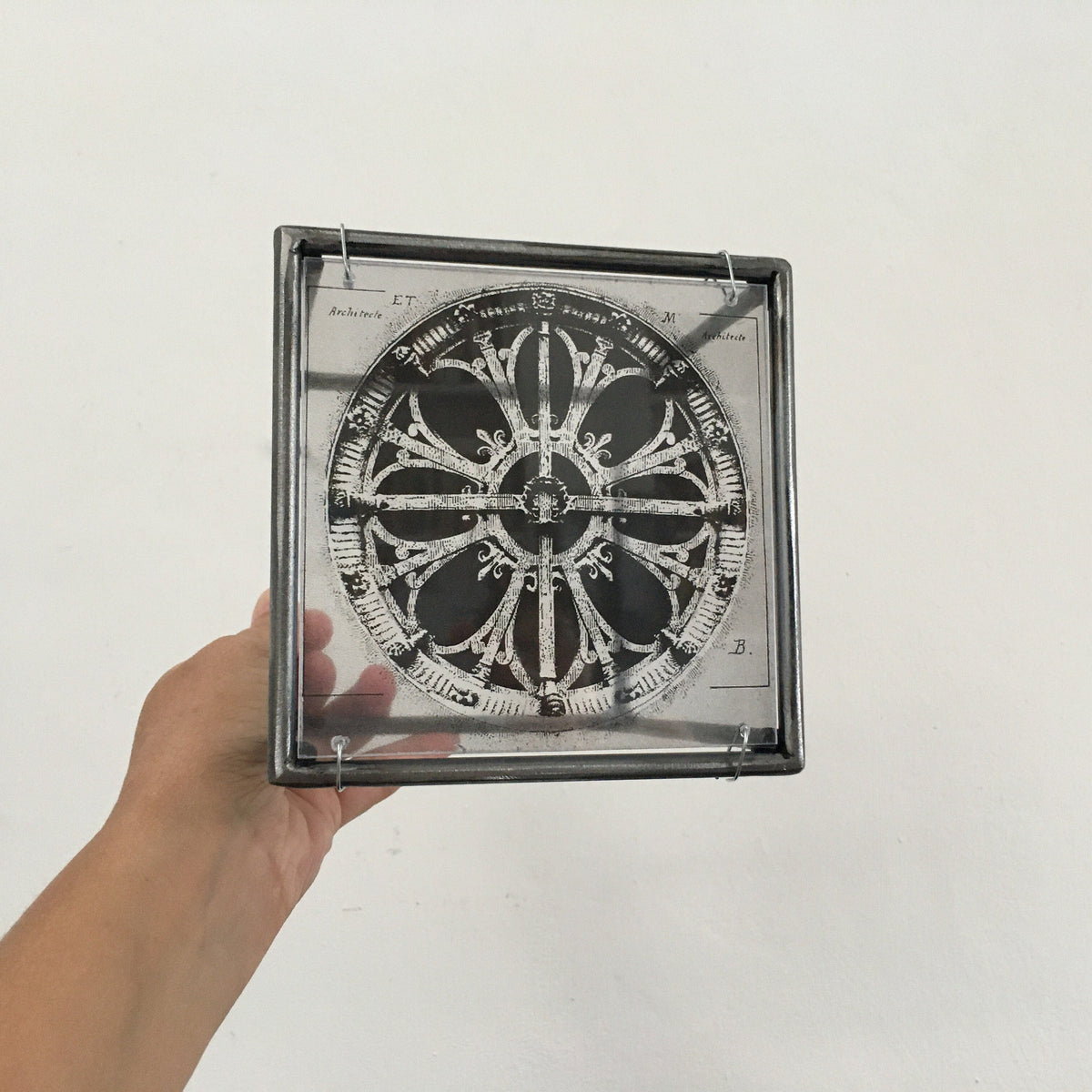 # human hand holding steel frame of vintage architectural drawings of two French rose windows —  Château de Chantilly and Église Saint-Léonard de Croissy-sur-Seine, printed on transparent acetate between acrylic glass. Modern Interior Metal Wall Art Sculpture Rose Window Diptych - Architectural wall art Artandshadow